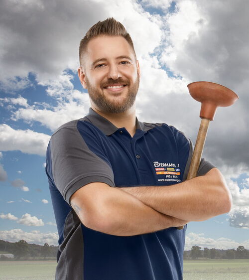 Armin Estermann jun. Handwerker mit blauem Shirt und WC-Pümpel vor Wiese und Wolkenhimmel