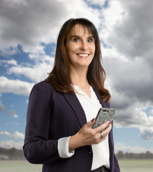 Dunkelhaarige Frau mit blauem Sakko, Mobiltelefon und weissem Hemd vor Wiese und Wolkenhimmel