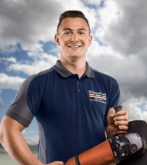 Handwerker mit blauem Shirt und Winkelschleifer vor Wiese und Wolkenhimmel