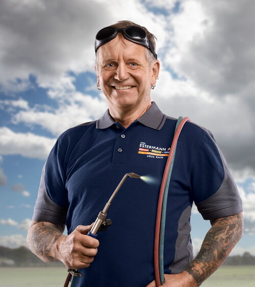 Handwerker mit blauem Shirt und Schweissgerät und Schweissbrille vor Wiese und Wolkenhimmel