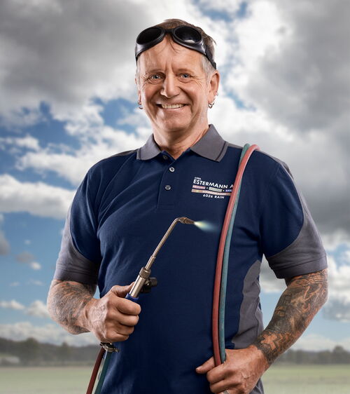 Handwerker mit blauem Shirt und Schweissgerät und Schweissbrille vor Wiese und Wolkenhimmel