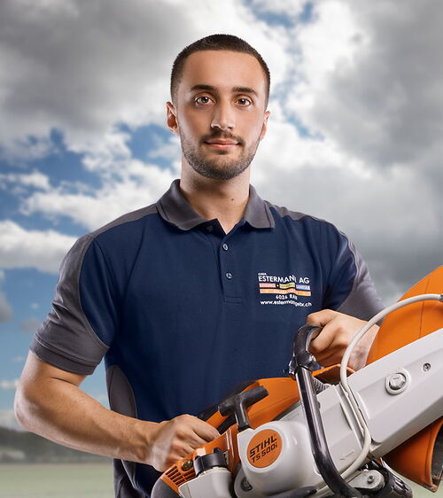 Raphael Odermatt - Sanitärinstallateur EFZ bei Gebr. Estermann AG Handwerker mit Bart, blauem T-Shirt und Trennsense vor Wolkenhintergrund