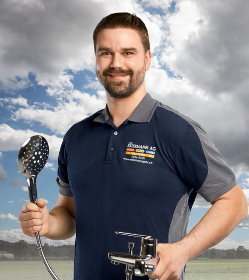 Martin Camenzind Handwerker mit blauem Shirt und Duschbrause und Mischbatteriel vor Wiese und Wolkenhimmel