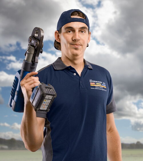 Handwerker mit blauem Shirt und Schweissgerät und Schweissbrille vor Wiese und Wolkenhimmel