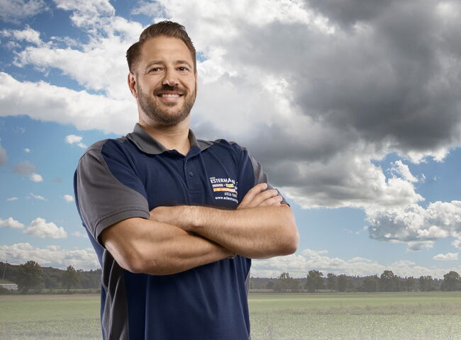 Handwerker mit blauem Shirt vor Wiese und Wald mit Wolken am blauem Himmel