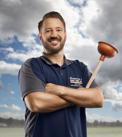 Handwerker mit blauem Shirt und WC-Pümpel vor Wiese und Wolkenhimmel