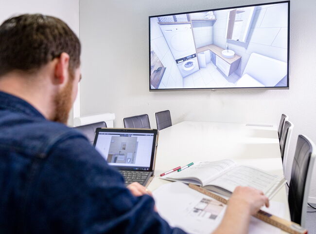 Mann mit blauem Hemd vor Laptop und Screen an weissem Tisch