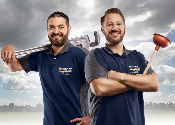 Zwei Handwerker mit blauem Shirt und Werkzeugen vor Wiese und Wald mit Wolken am blauem Himmel